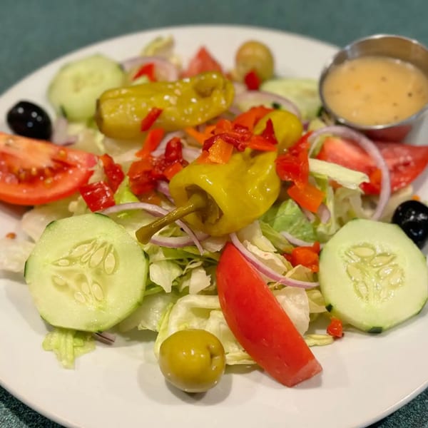 A fresh Italian salad with lettuce, tomatoes, cucumbers, olives, onions, and peppers, with dressing on the side.
